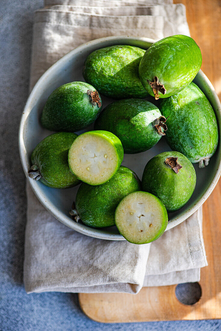Schale mit reifen Feijoa-Früchten auf einer hölzernen Oberfläche, mit einem rustikalen Tuch, das die gesamte grüne Feijoa und ihren Querschnitt zeigt
