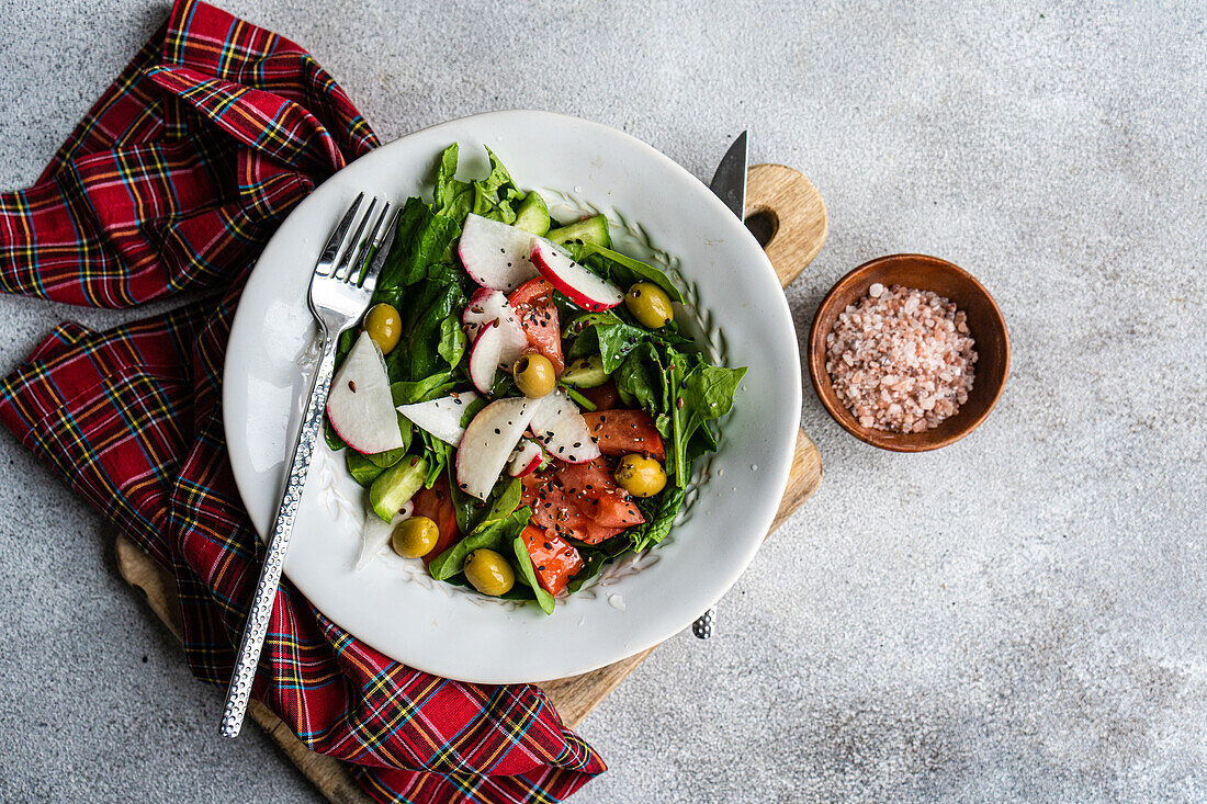 A vegetable salad made with fresh organic spinach leaves, sliced tomatoes, radishes, and olives presented in a white bowl beside a plaid cloth, wooden cutting board, and a bowl of pink salt