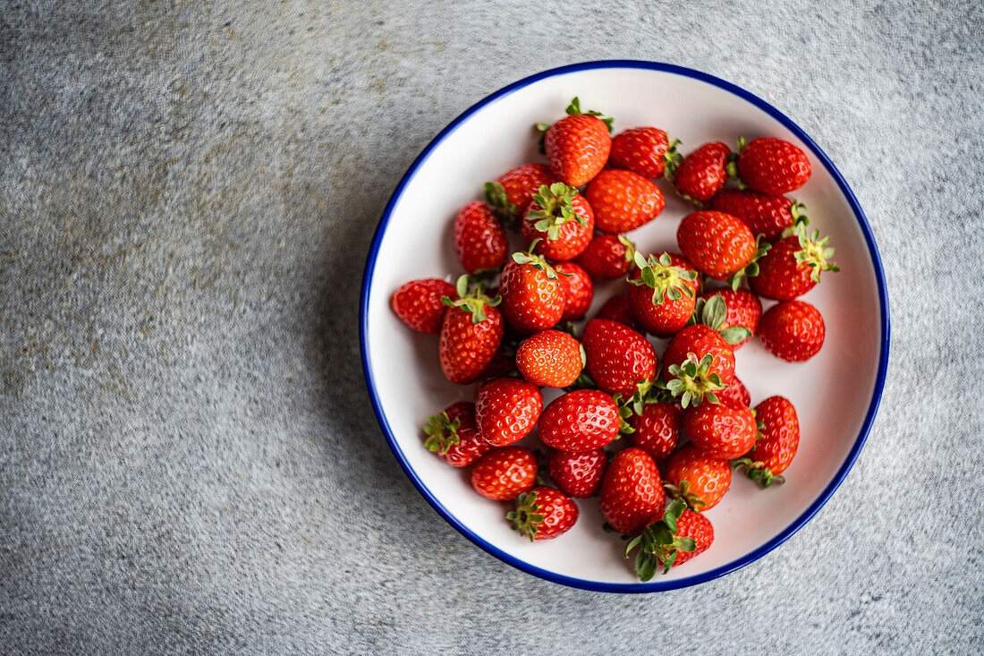 Top view of bright red organic strawberries displayed on a white plate with a blue rim, placed against a textured grey background.