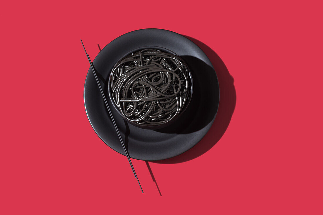 Draufsicht auf schwarze Spaghetti in einer Schüssel mit Stäbchen auf schwarzem und rotem Hintergrund
