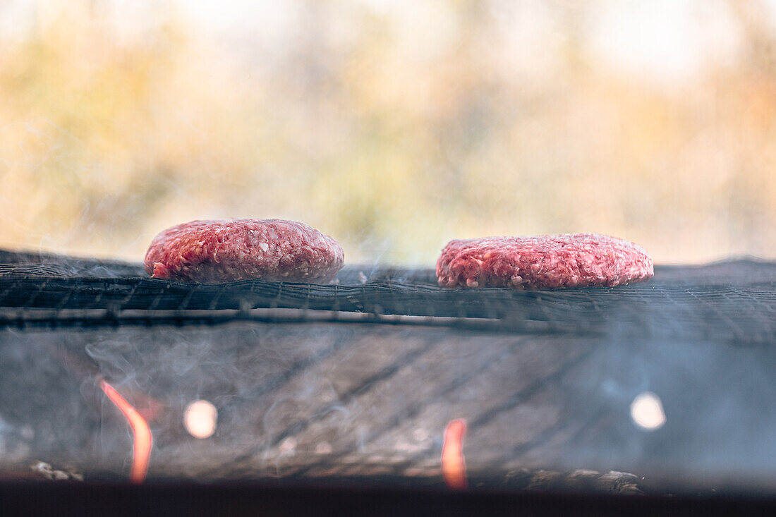 Two burger patties smoke enticingly on a grill with glowing embers beneath