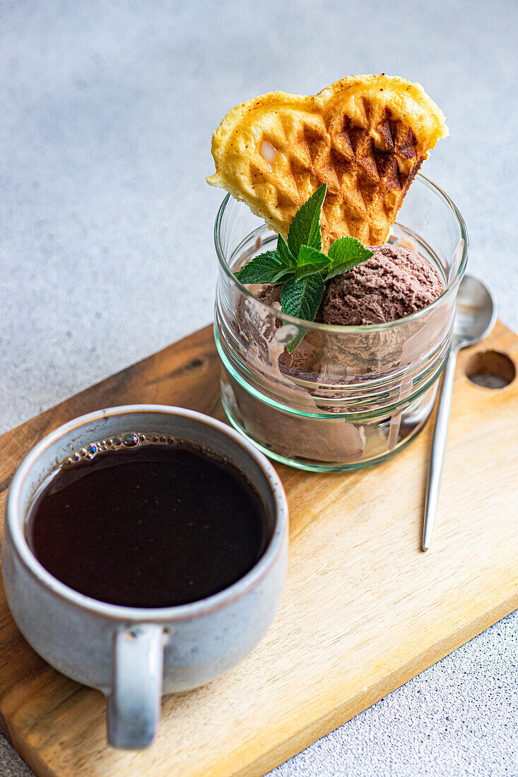 High angle of glass of homemade coffee ice cream with waffle and fresh mint near spoon and cup of coffee placed on wooden tray against gray surface