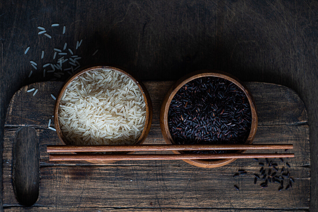 Top view of Raw wild black rice and peeled white rice in the bowls with chopsticks placed on cutting board on wooden table