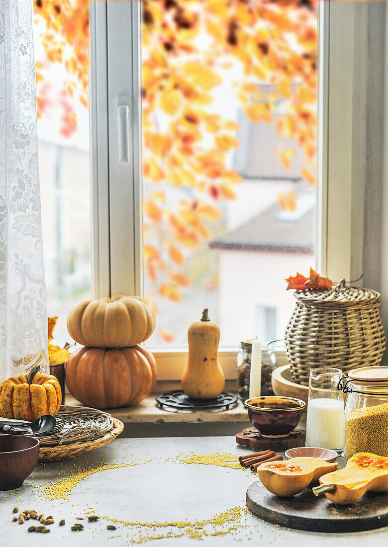 Herbstliches Küchenstillleben mit verschiedenen Kürbissen, Küchenutensilien und Zutaten am Betontisch mit Fensterhintergrund mit Herbstlaub