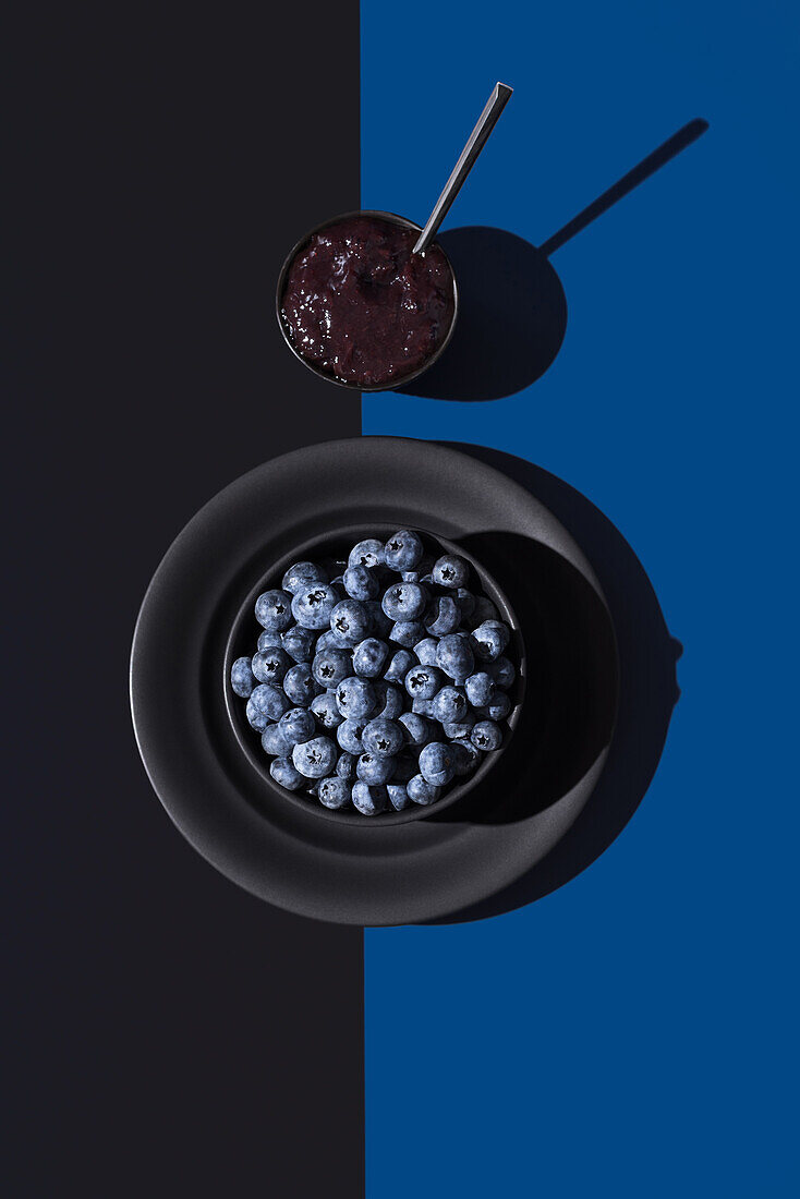 Fresh blueberries in a black bowl with a cup of blueberry jam on a split blue and black background, showcasing a play of light and shadow