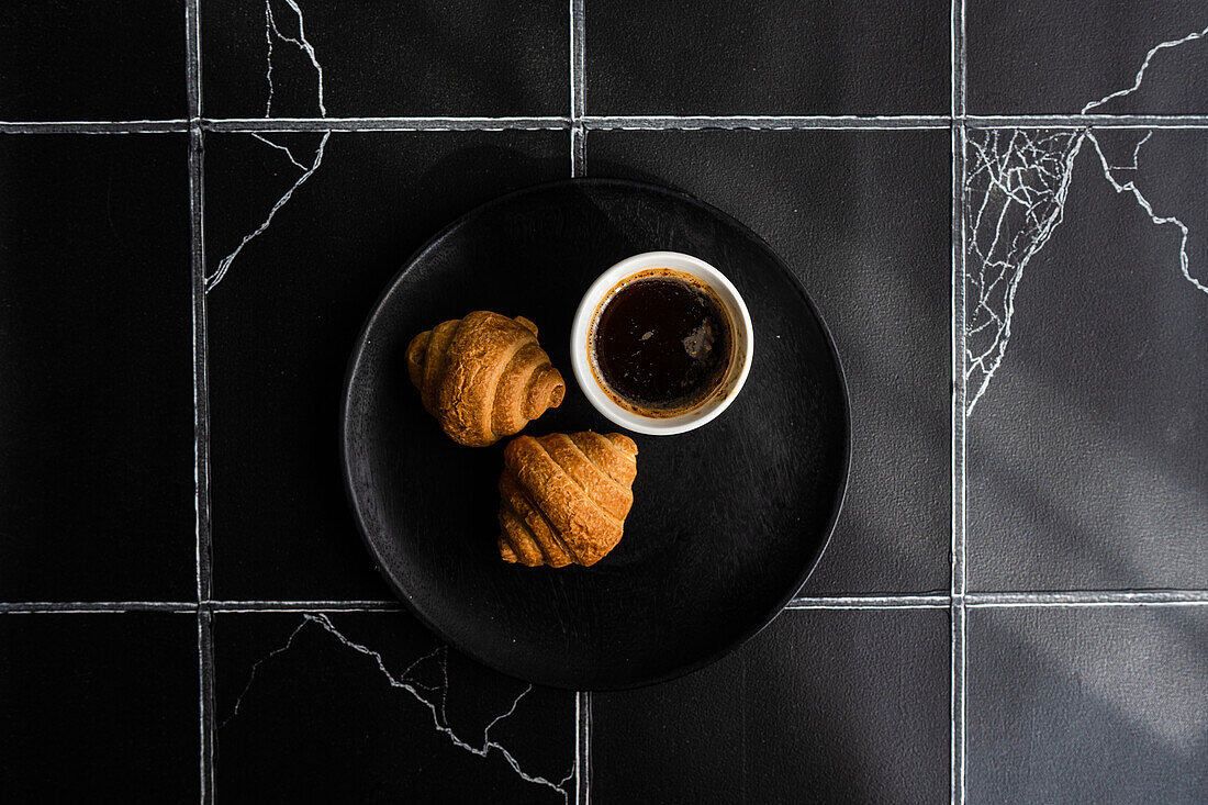 Draufsicht auf einen schwarzen Kaffee und frisch gebackene Croissants auf einem schwarzen Betontisch