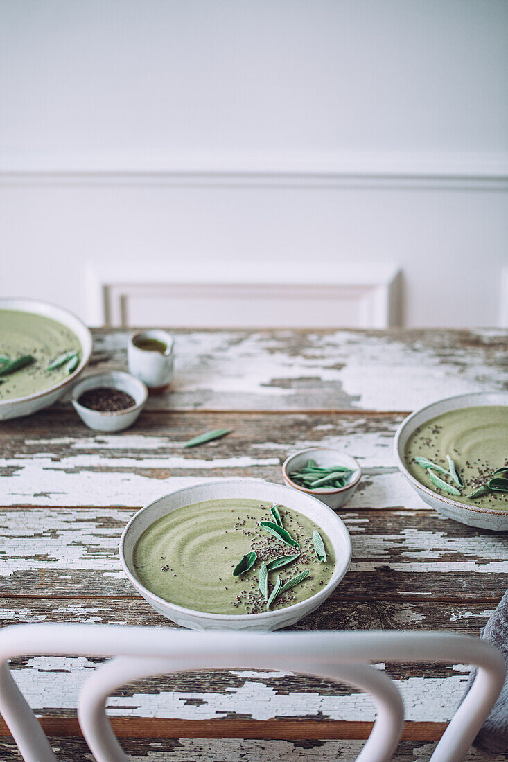 Keramikschalen mit grüner Cremesuppe mit Salbei, serviert mit Samen und Kräutern auf einem Holztisch mit schäbiger Oberfläche vor weißem Hintergrund