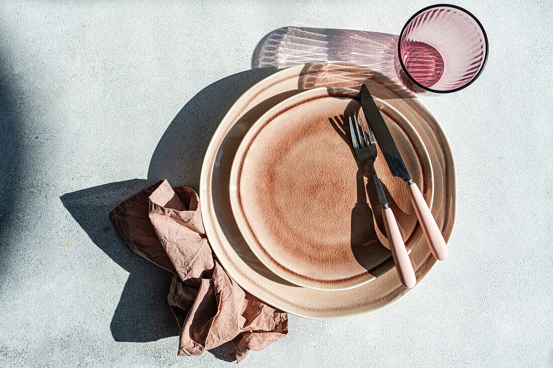 Von oben: Raffiniert gedeckter Ostertisch mit gestapelten Keramiktellern, roségoldenem Besteck und einer Leinenserviette vor einem strukturierten Hintergrund