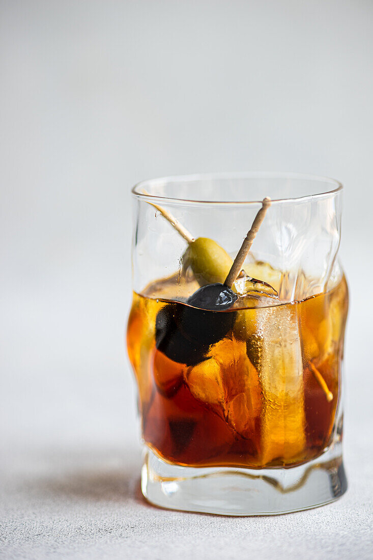 Transparent glass cup with whiskey alcoholic drink and stick of olives cocktail with ice cubes on grey background in studio