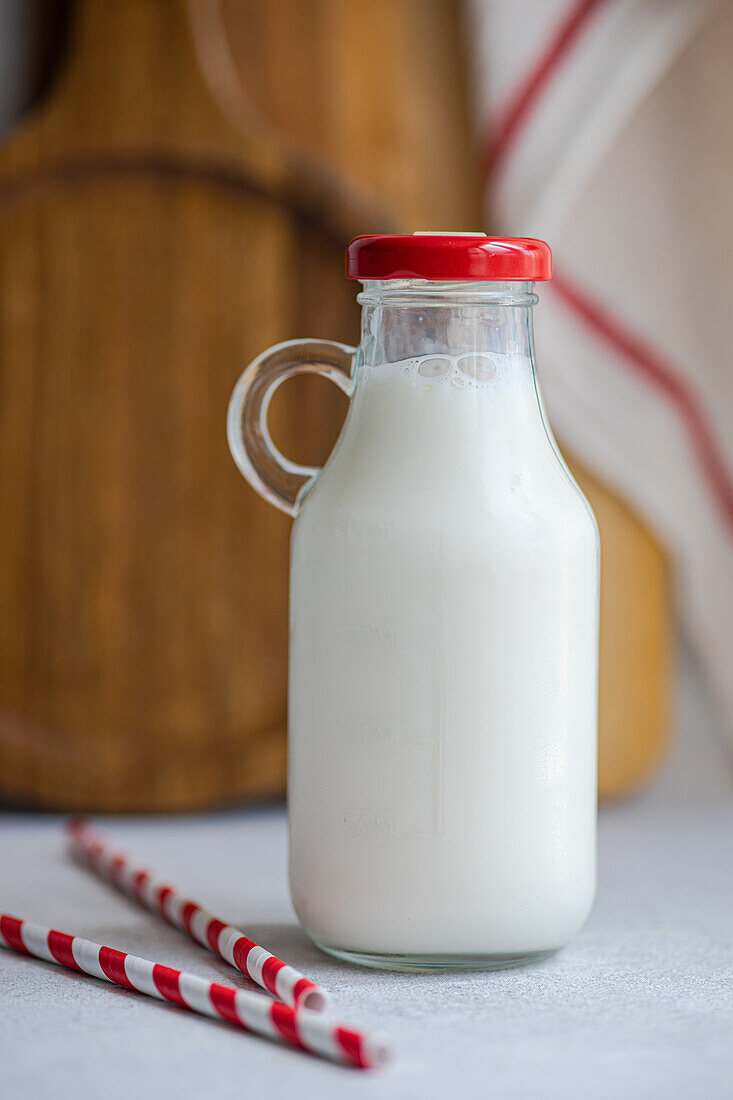 Rohe Kuhmilch in verschlossener Flasche neben Trinkhalmen auf grauer Fläche vor unscharfem Hintergrund