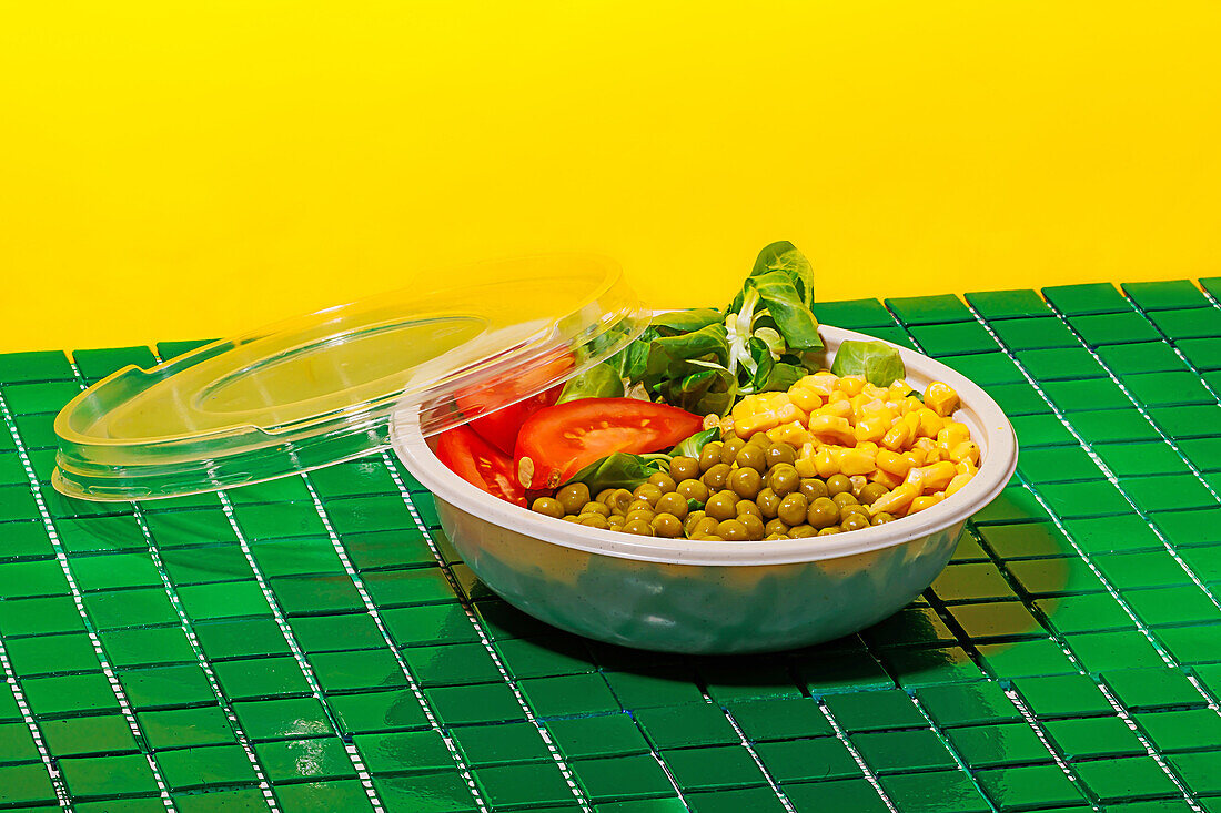 Salad bowl with slices of tomato, spinach leaves, corn kernels and peas placed on green surface against yellow wall