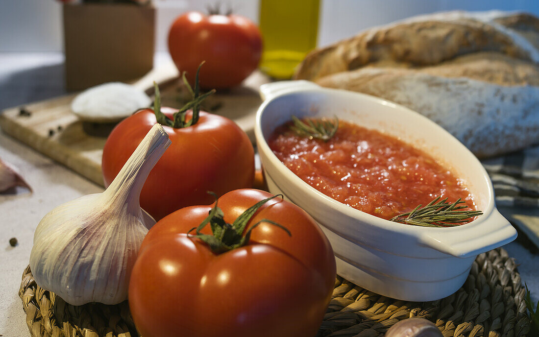 Close up of ripe tomatoes and unpeeled garlic placed on table near bowl with crushed tomatoes, wholegrain bread on napkin, chopping board and oil