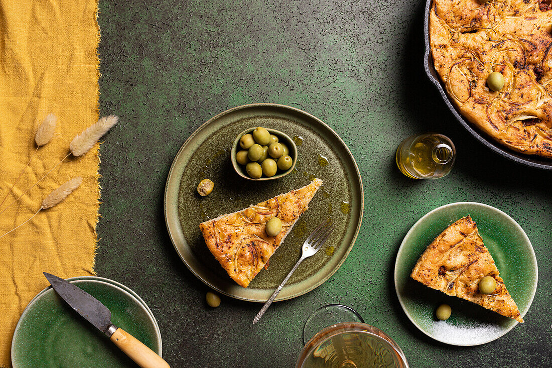 Draufsicht auf eine Scheibe gebackenes italienisches Focaccia-Brot mit grünen Oliven in einem Teller auf einem Esstisch mit Getränk auf grünem Hintergrund