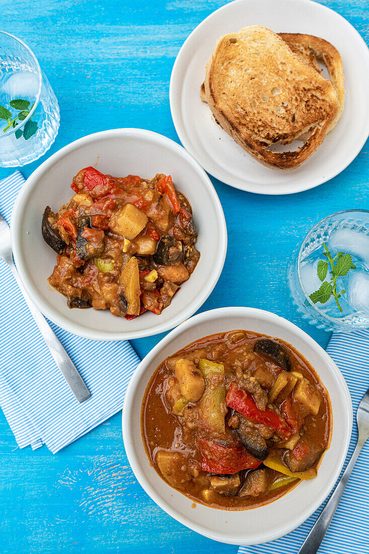 Top view of traditional seasonal Georgian dish - Adjapsandali with stewed vegetables like potato, bell peppers, tomato, egg plant, coriander and Georgian spices placed on blue table near napkin and glass with water