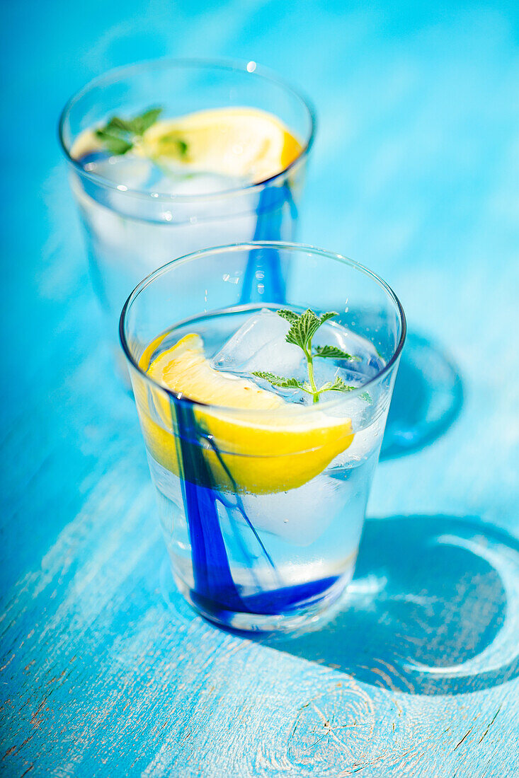 Hochformatiger Sommercocktail mit Zitronenwodka, Zitronenscheiben und Minzblättern, serviert mit Eis auf einem blauen Tisch