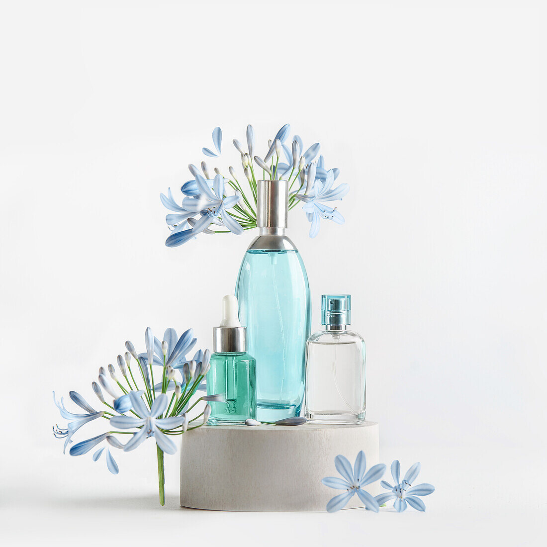 Schönheitsprodukte in blauen Glassprüh- und Pipettenflaschen stehen auf einem Podest mit Blumen und Blütenblättern vor weißem Hintergrund. Kosmetisches Setting mit Serum, Parfüm, Duft und Toner. Vorderansicht