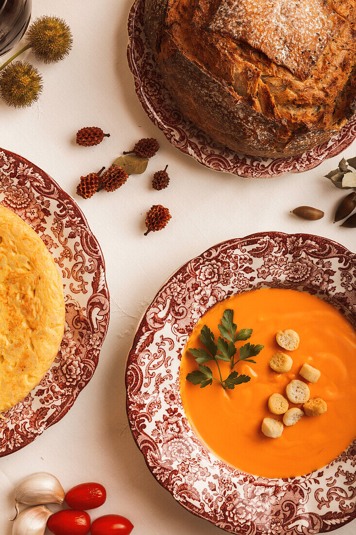 A cozy autumnal spread showcasing a tasty spanish omelette and a creamy salmorejo soup wit croutons and bread on decorative plates.
