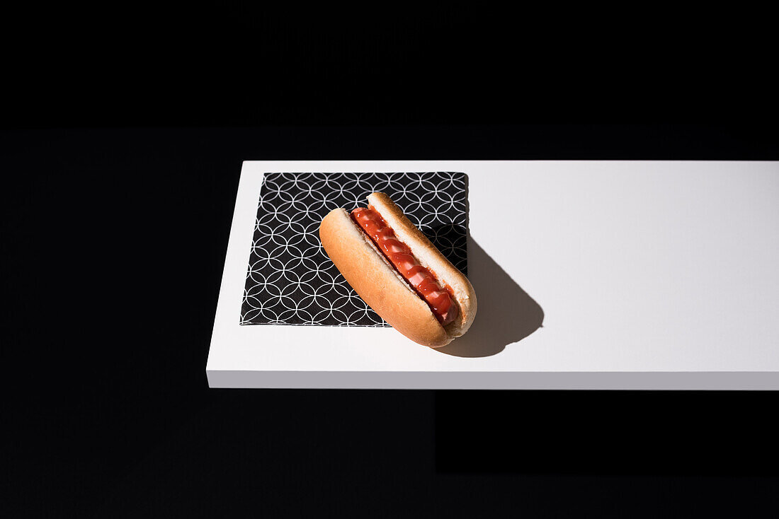 Blick von oben auf ein appetitliches Brötchen mit Wurst und Ketchup, serviert auf einem weißen Holzbrett vor schwarzem Hintergrund im Studio