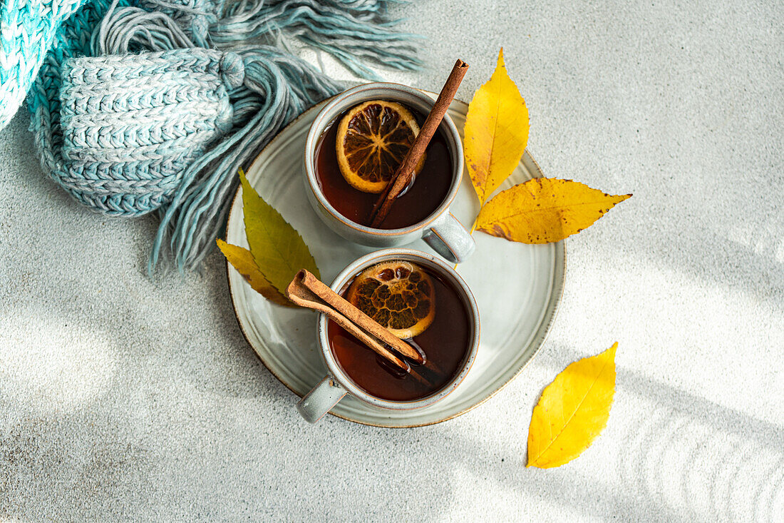Top view of cups of spiced tea adorned with cinnamon sticks, anise, and dried orange slices, nestled next to vibrant yellow leaves and a folded blue scarf on a soft gray backdrop