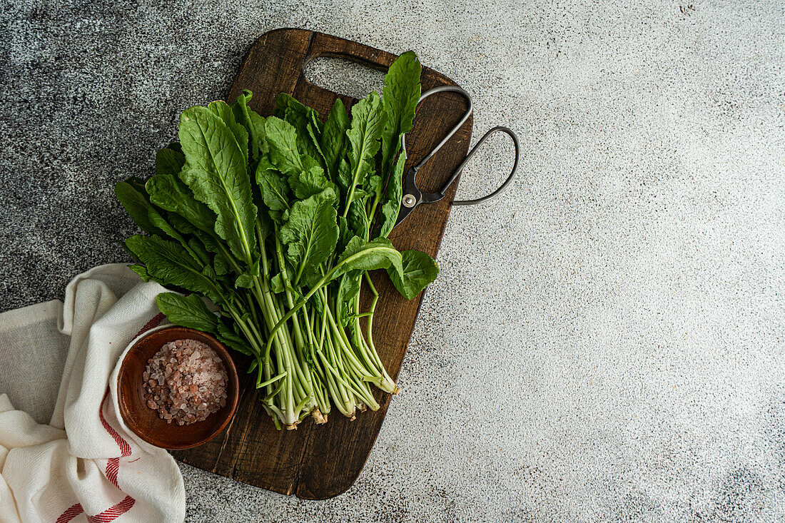 Top view of bunch of fresh arugula herb leaves on a rustic wooden cutting board, accompanied by a bowl of pink salt and kitchen shears, set on a grey textured surface