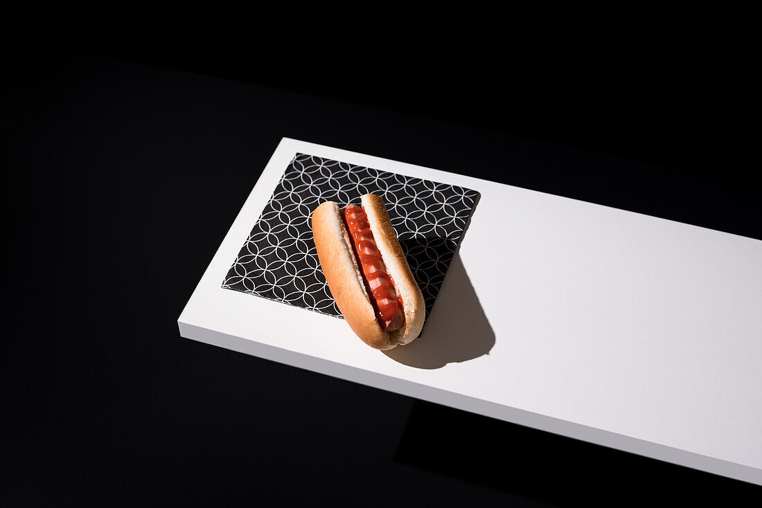 High angle of appetizing bun with sausage and ketchup served table mat over white wooden board against black background in studio