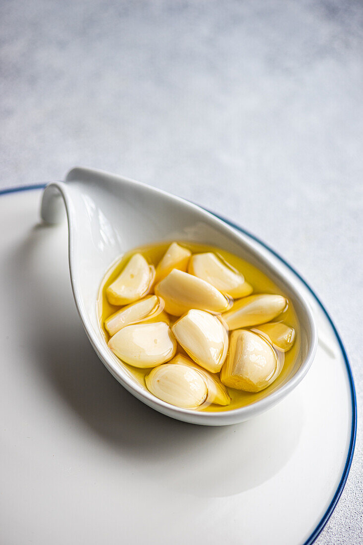 High angle of peeled and baked garlic cloves submerged in oil, showcased in a ceramic spoon placed on a white plate with a subtle blue border, against a light gray background