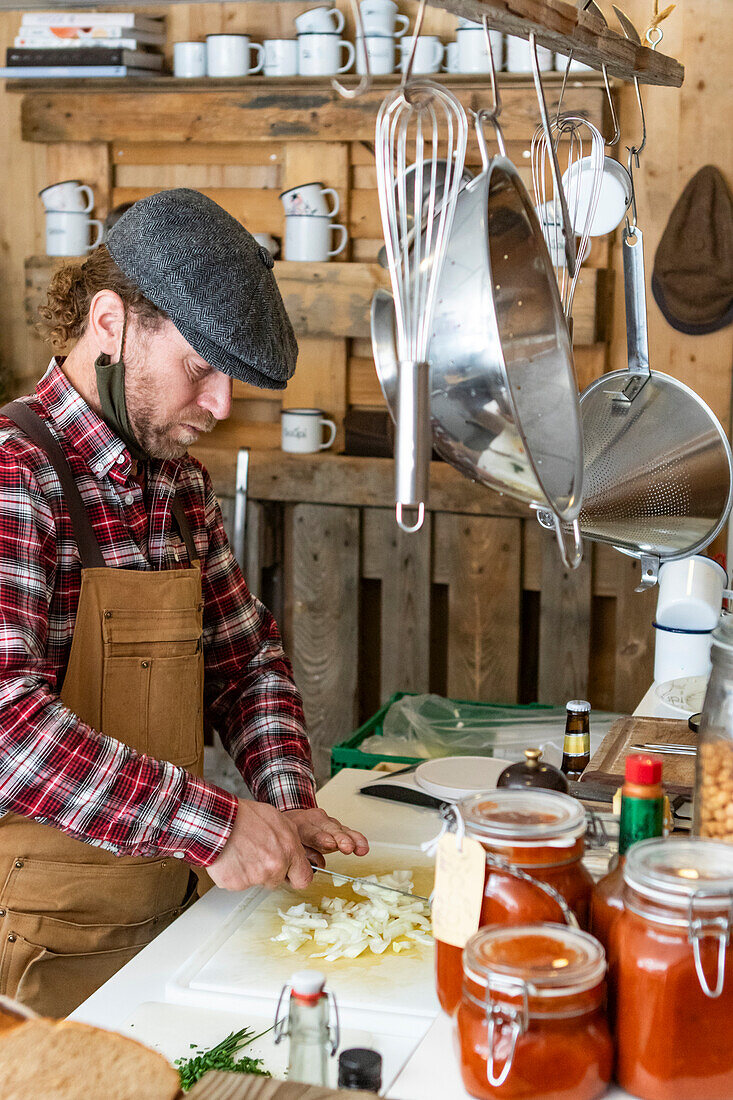 Side view of mature focused chef in apron and beret looking down while chopping onions, surrounded by a rustic setting with hanging kitchenware and jars of homemade preserves