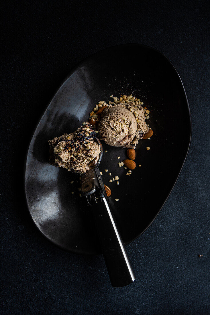 Draufsicht auf einen schwarzen Keramikteller mit einer Kugel Eis, der neben einer Kugel Schokoladeneis mit Erdnüssen und Walnüssen steht