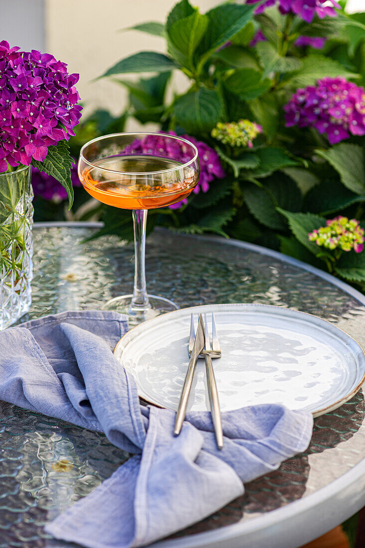 Vase mit lila Hortensien steht auf dem Tisch neben einem Glas mit Getränk und einem Keramikteller im Garten