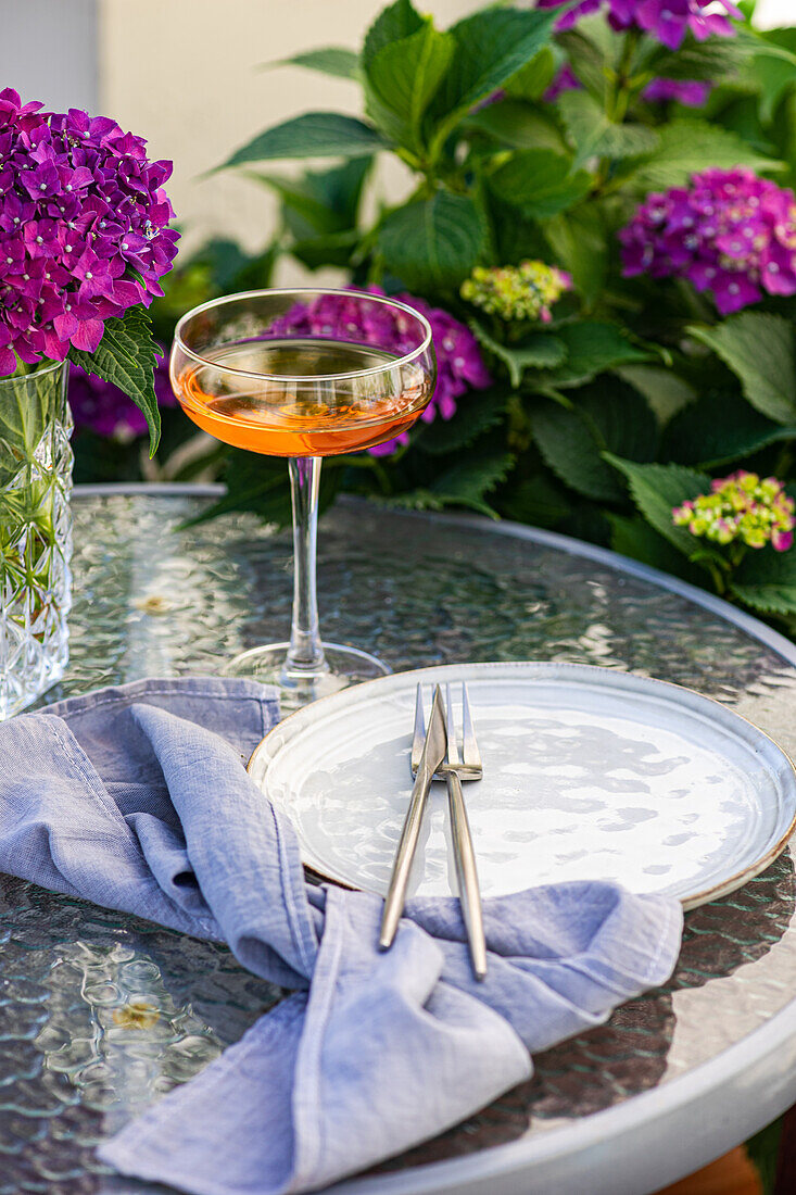 Vase with purple hydrangeas places on table near glass with drink and ceramic plate in garden