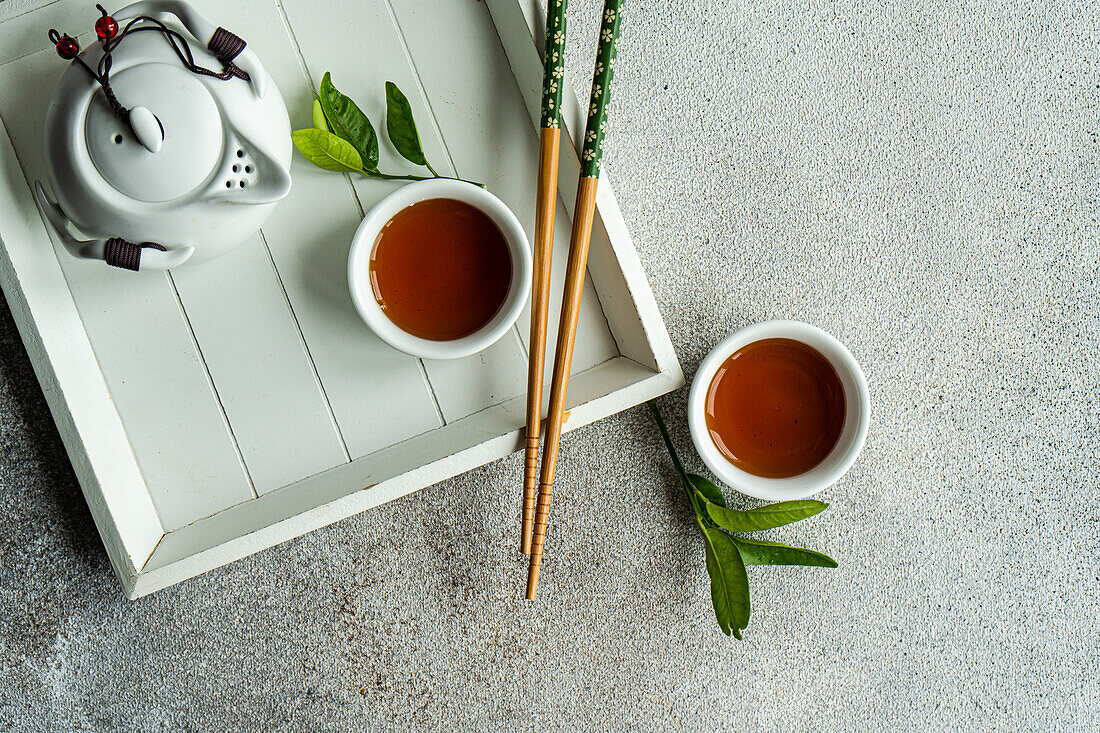 Draufsicht auf Ernte-Tee-Set im asiatischen Stil mit Zitronenblättern und Stäbchen auf weißem Tablett vor grauem Hintergrund