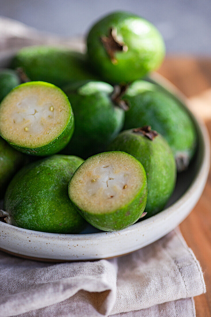 A close-up view of fresh feijoa fruits, with one sliced in half, in a ceramic bowl on a linen cloth.