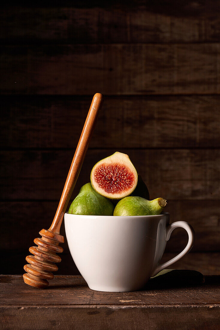 A rustic still life of fresh green figs, one sliced open to showcase the red interior, paired with a wooden honey dipper, all beautifully arranged in a white ceramic cup set against a dark wooden backdrop.