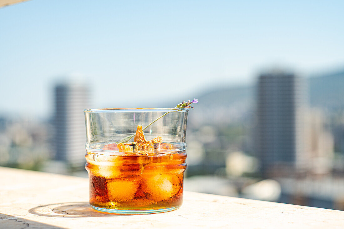 Glas Whiskey mit Eis, Orangenschale und Blume auf weißer Fläche vor unscharfem Gebäude