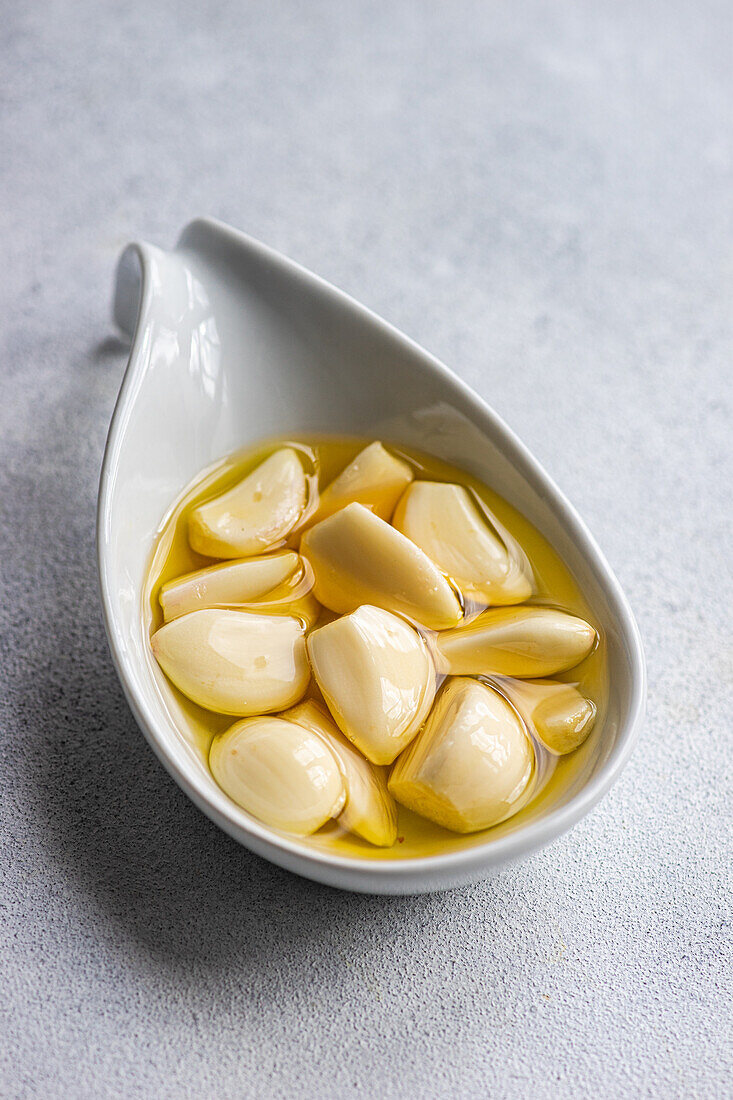 High angle of peeled and baked garlic cloves submerged in oil, showcased in a ceramic spoon against a light gray background