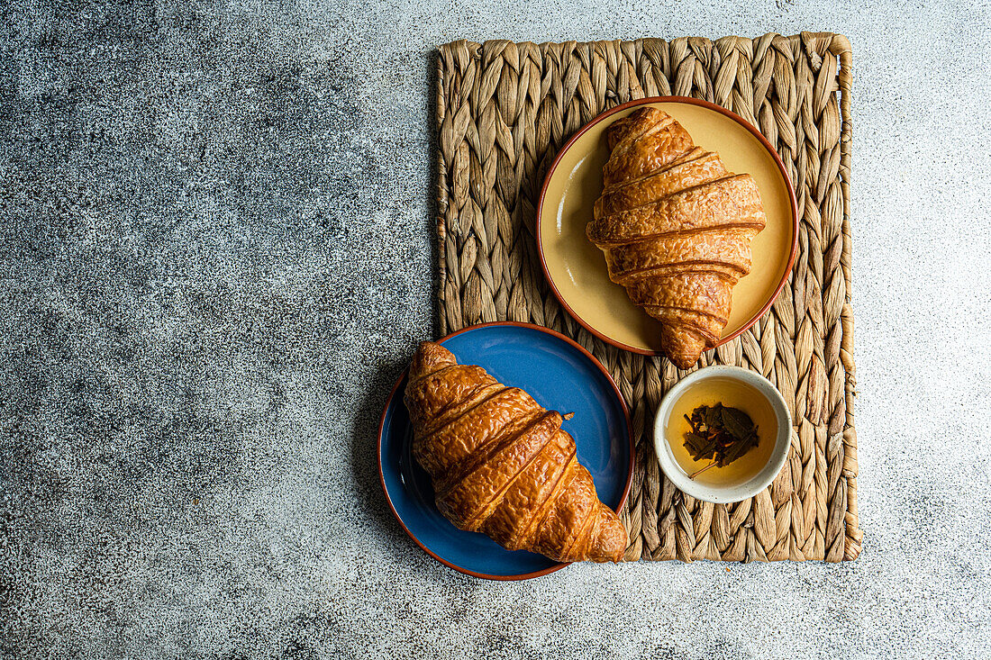 Draufsicht auf frisch gebackene Croissants auf bunten Keramiktellern auf brauner Serviette neben einer Tasse grünem Tee vor grauem Hintergrund