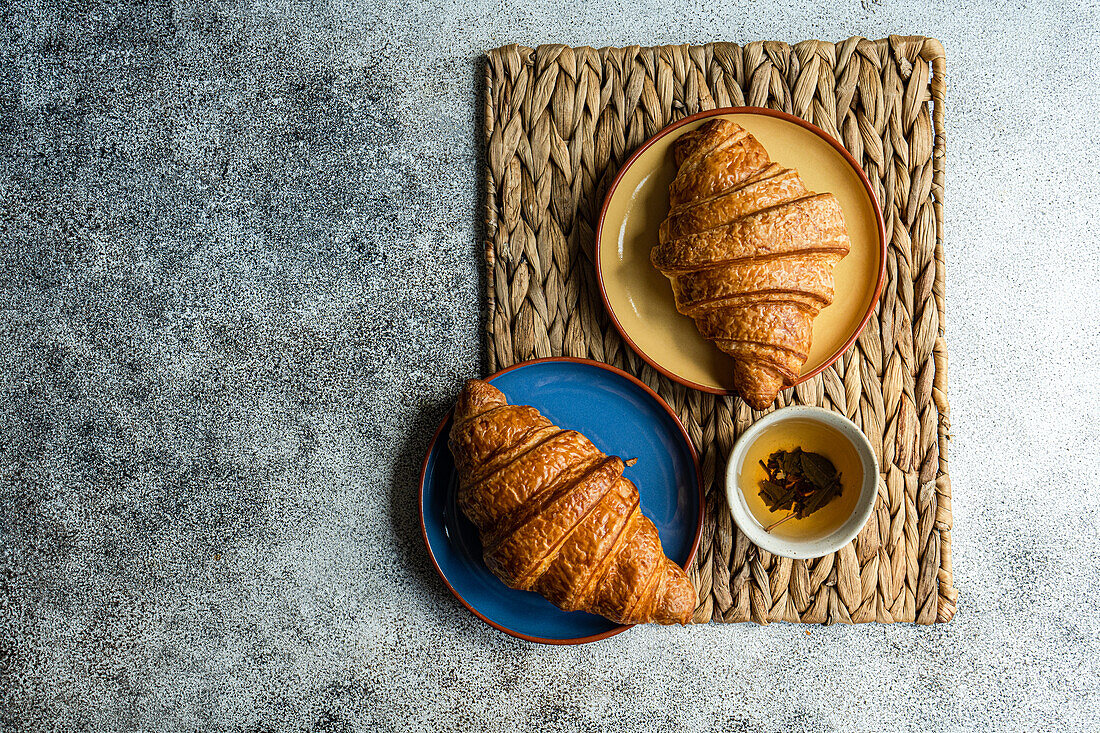 Top view of fresh baked croissants on the colorful ceramic plates placed on brown napkin near cup of green tea against gray background