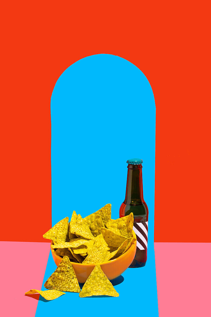 A vibrant still life image featuring a bowl of crunchy nacho chips beside a dark bottle against a split background of red and blue