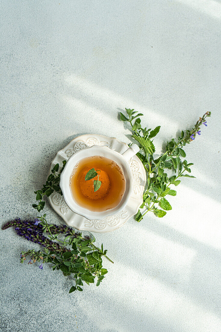 Draufsicht auf ein Glas Blumentee mit frischen Kräutern, serviert auf einem Keramikteller vor grauem Hintergrund