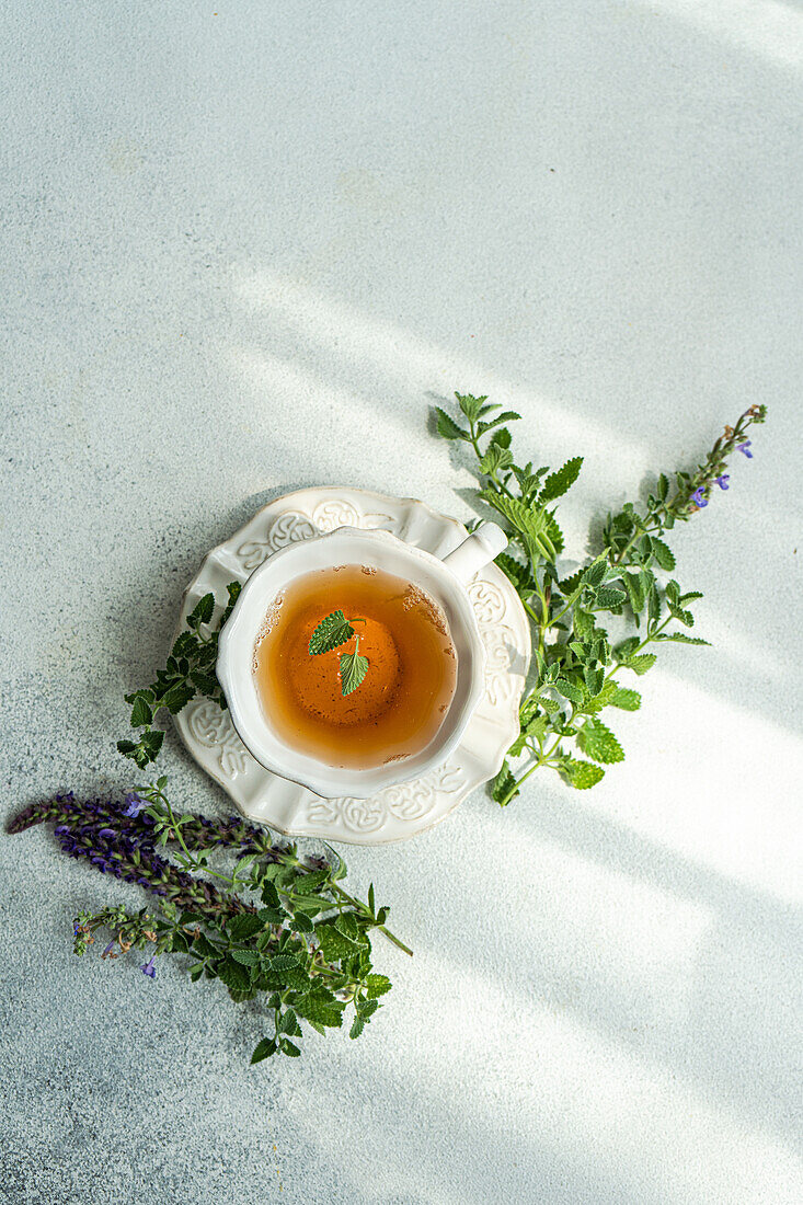 Top view of glass of floral tea with fresh herbs served on ceramic plate against gray background