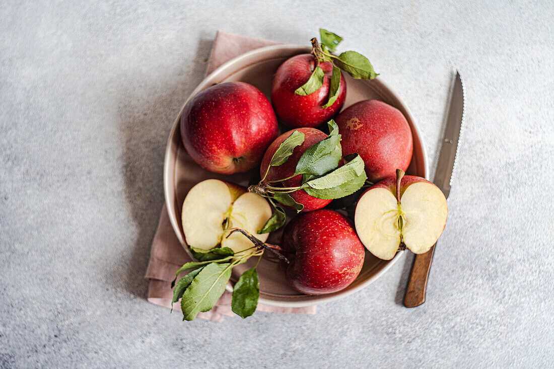 Draufsicht auf einen Strauß reifer roter Äpfel in einem Teller neben einem Messer und einem Tuch auf einer grauen Fläche vor einem unscharfen Hintergrund