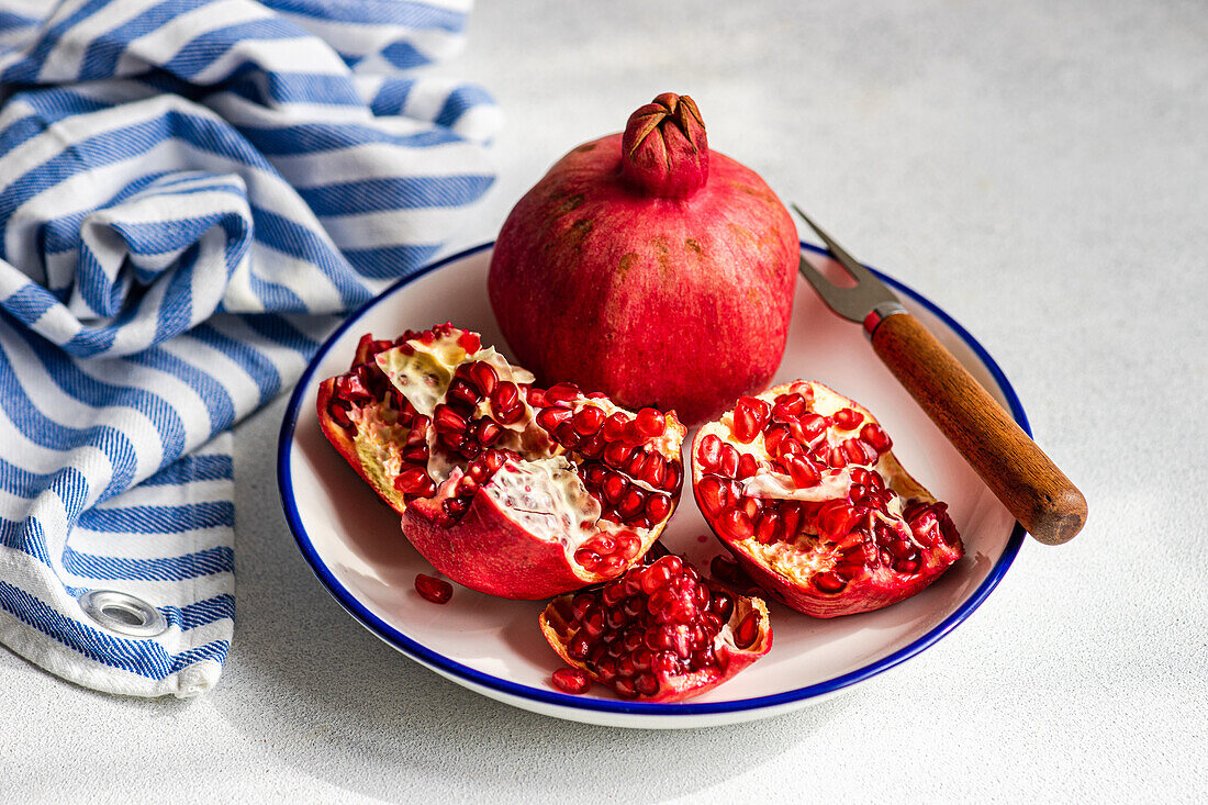 A fresh ripe pomegranate sits on a white and blue plate, surrounded by its red seeds and a rustic knife with a wooden handle. A striped napkin blue and white against gray background.