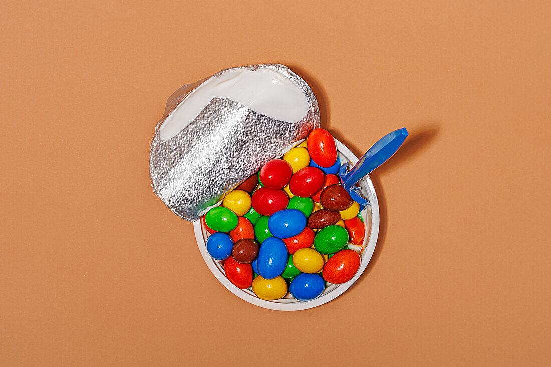 Top view of opened yoghurt container filled with colored candies and spoon against orange background