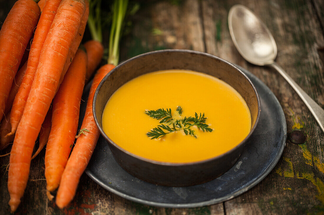 A bowl of creamy carrot soup garnished with parsley alongside raw carrots and a vintage spoon on a rustic wooden table.