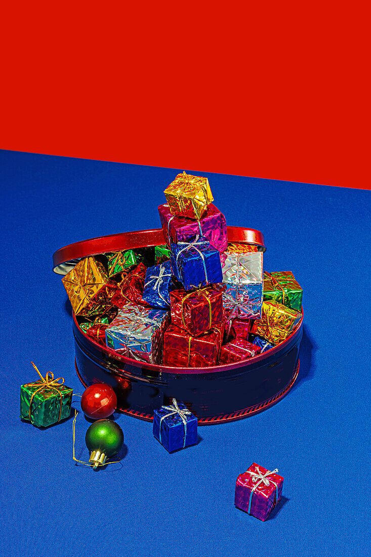 A blue tin filled with an assortment of shiny wrapped Christmas presents on a blue surface with a red and blue background