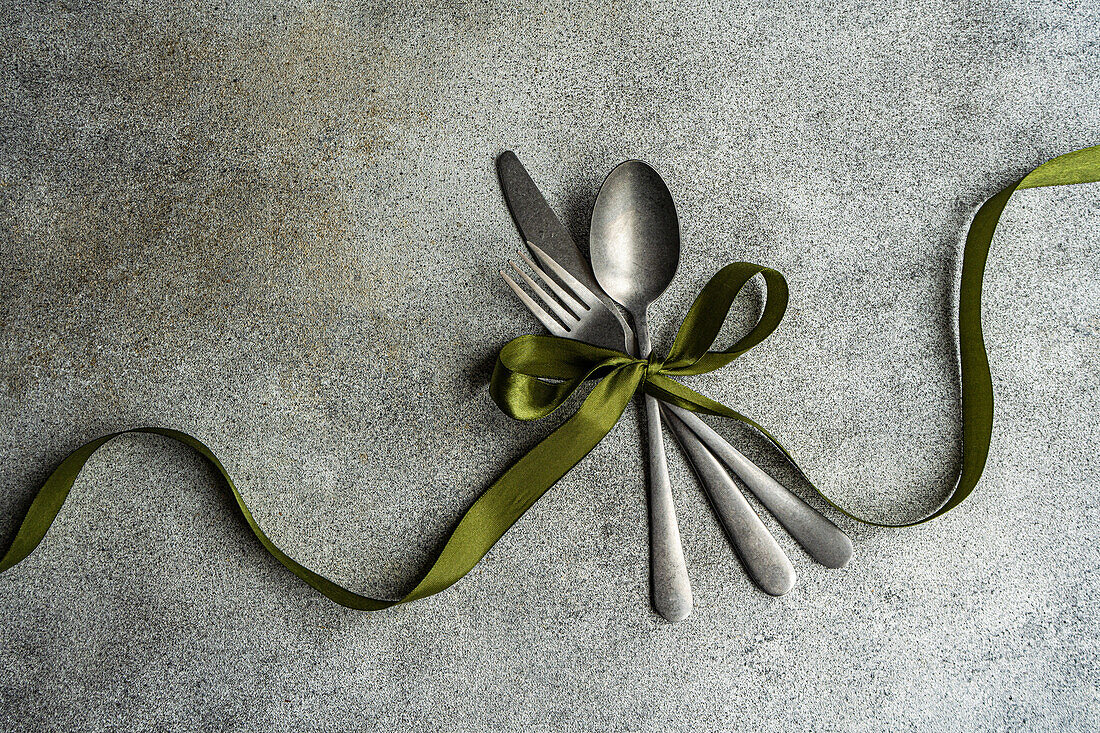 Top view of vintage cutlery set placed on gray surface with green ribbon in light kitchen
