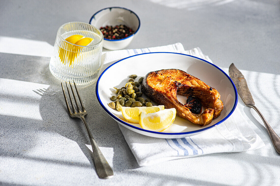 High angle of well-prepared grilled trout steak with capers and lemon served on a white plate with a blue rim placed on striped napkin, cutlery, glass of water and bowl with capers
