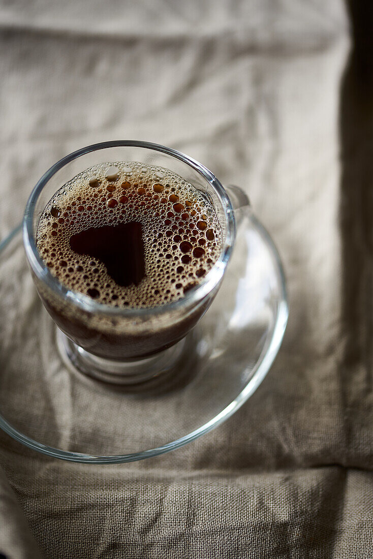 A detailed close-up shot capturing the frothy texture of freshly brewed coffee in a transparent glass cup set against a textured fabric backdrop