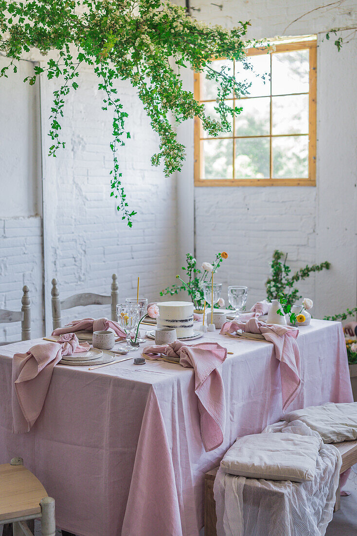 Eleganter Tisch mit Tellern und Blumen neben einem leckeren Kuchen auf einer rosa Tischdecke neben einer mit einem Tuch bedeckten Bank vor einer Topfpflanze und einer weißen Ziegelwand