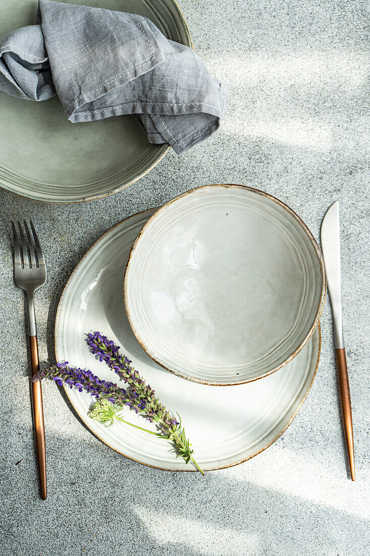 Draufsicht auf einen herbstlich gedeckten Tisch mit Keramikschüssel und Teller mit Lavendelblüten zwischen Gabel und Messer vor grauem Hintergrund